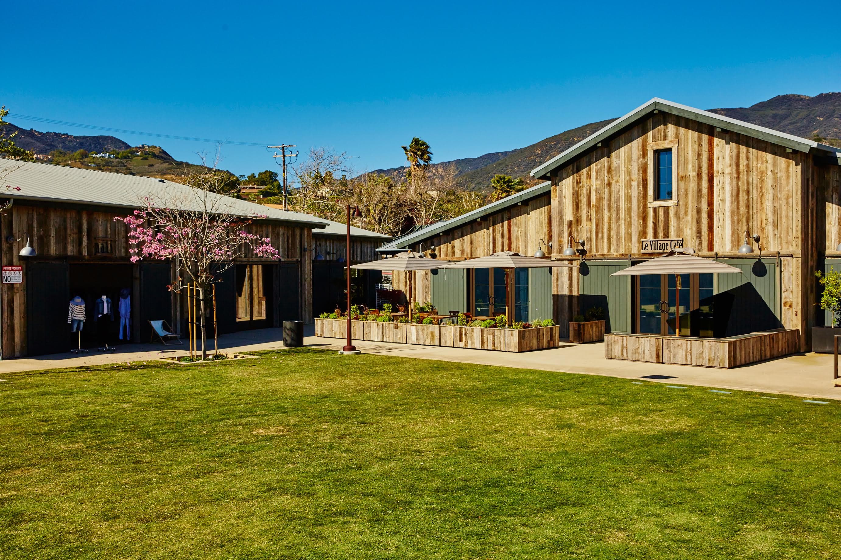 Daytime courtyard at Trancas Country Market: weathered wood barns, umbrellas, and a lawn under a clear Malibu sky. The building still shows the former Le Village Cafe name.